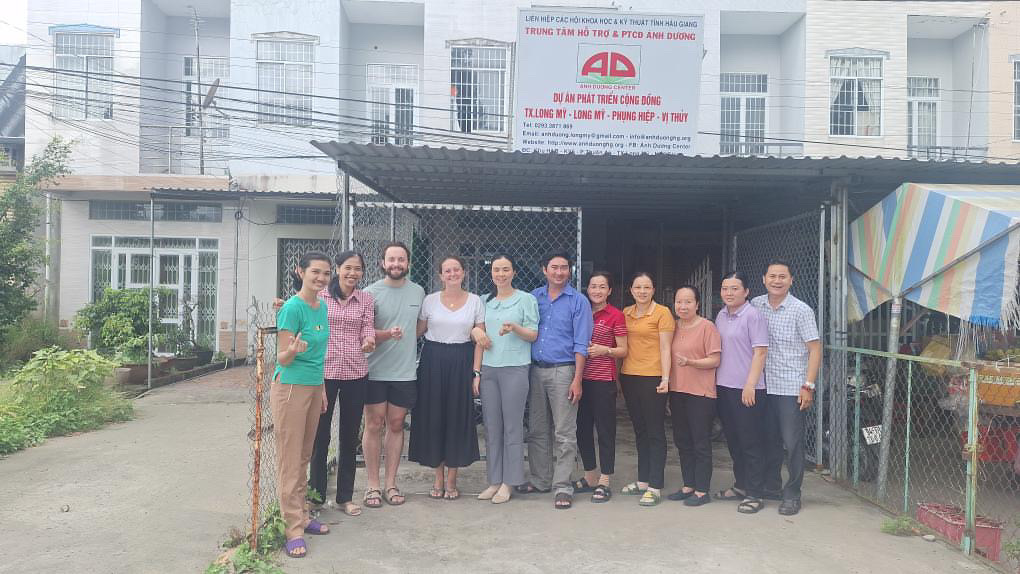 Hai tình nguyện viên chụp ảnh cùng cán bộ Trung tâm Ánh Dương trước văn phòng, dưới biển hiệu Trung tâm Hỗ trợ và Phát triển cộng đồng Ánh Dương